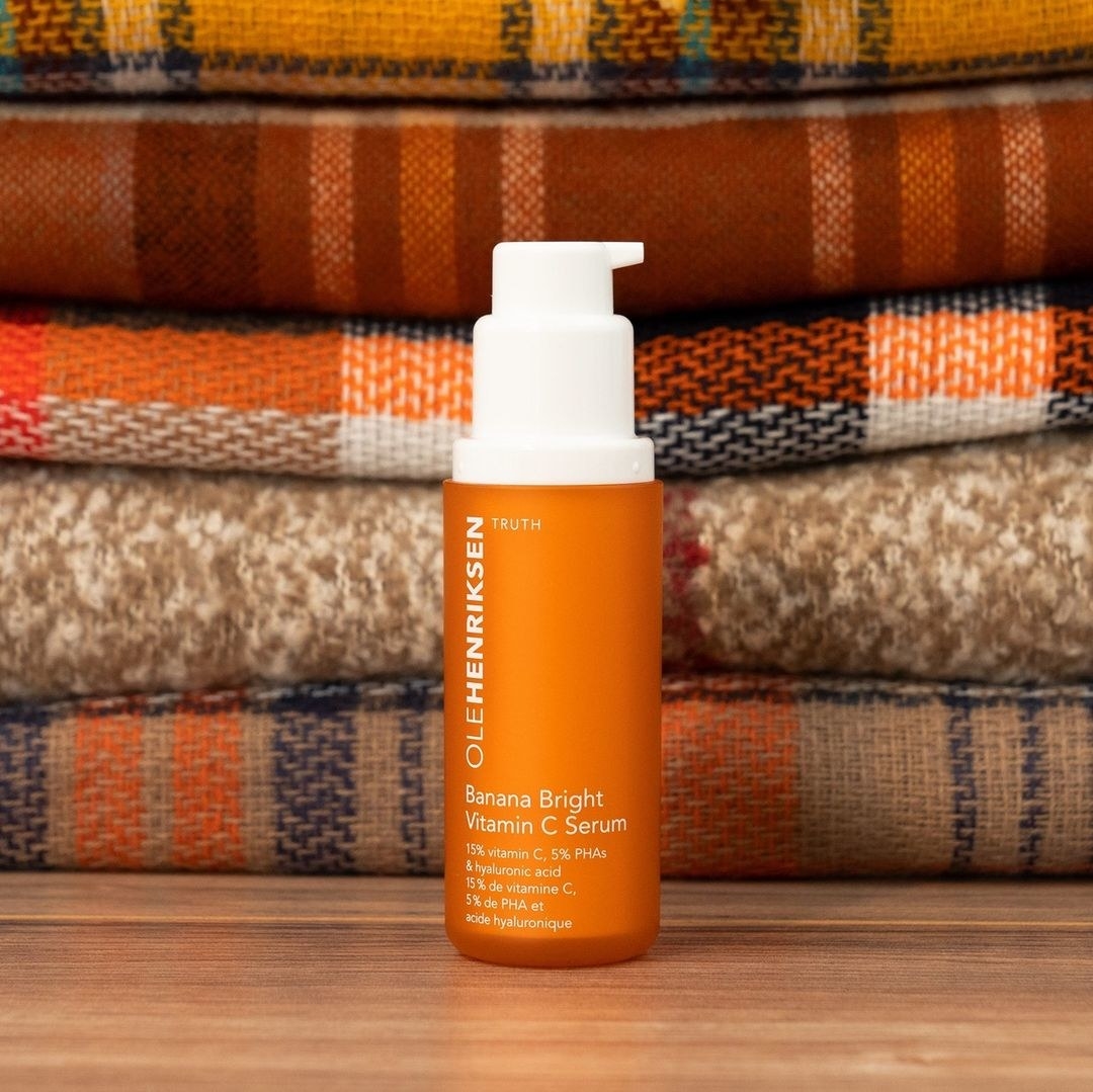A bottle of face serum on a wooden table with a stack of folded blankets behind it