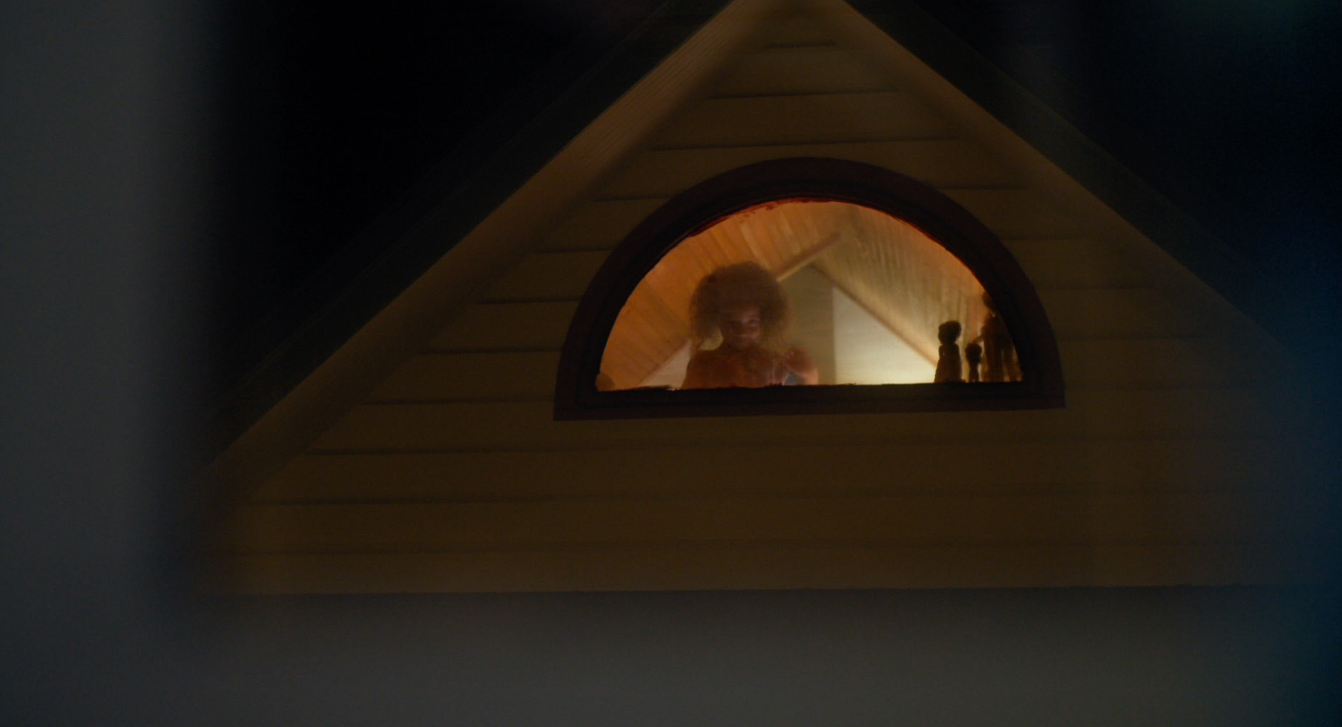 A small child stands in the half-circle window of an attic. It&#x27;s dark out but the light&#x27;s on in the attic.