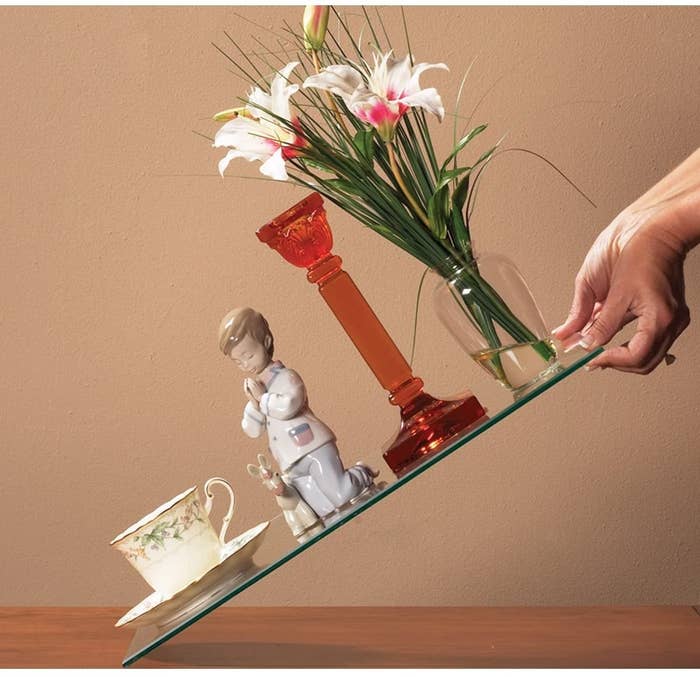 person holding a piece of glass at an angle that has a teacup, a porcelain doll, a candle holder, and a vase all secured so they don&#x27;t fall