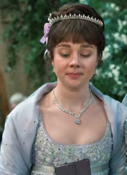 Eloise wears a blue gown and shawl with a tiara and purple bow
