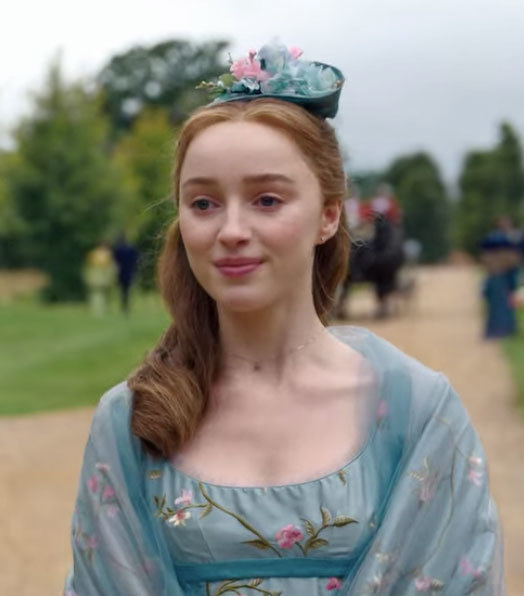 Daphne wears a blue dress embroidered with flowers and a matching shall and hat