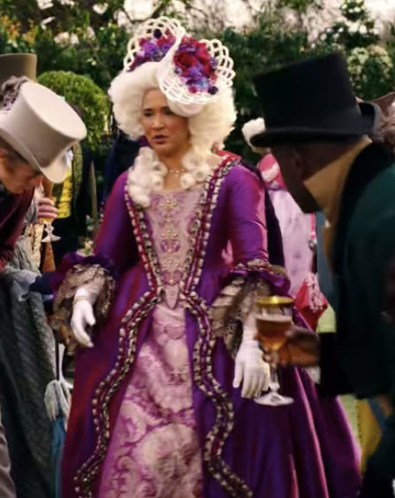 The queen wears an elaborate purple dress and headpiece