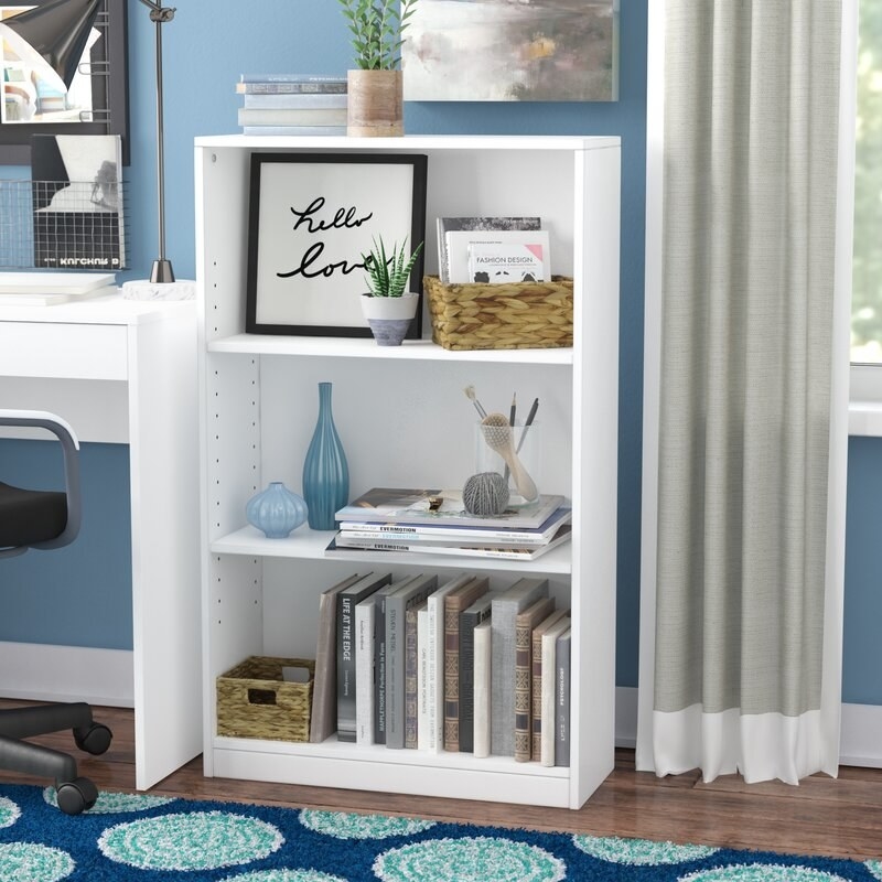 a white bookcase with three shelves, holding books, art and decor in a room