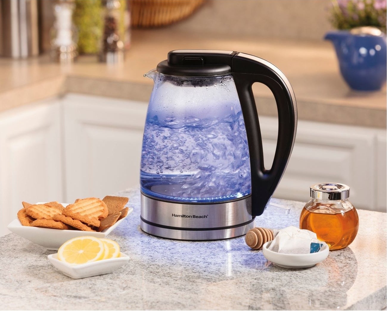 Glass kettle with blue light, stainless steel base and black handle sits beside yellow lemons, tan cookies and amber honey