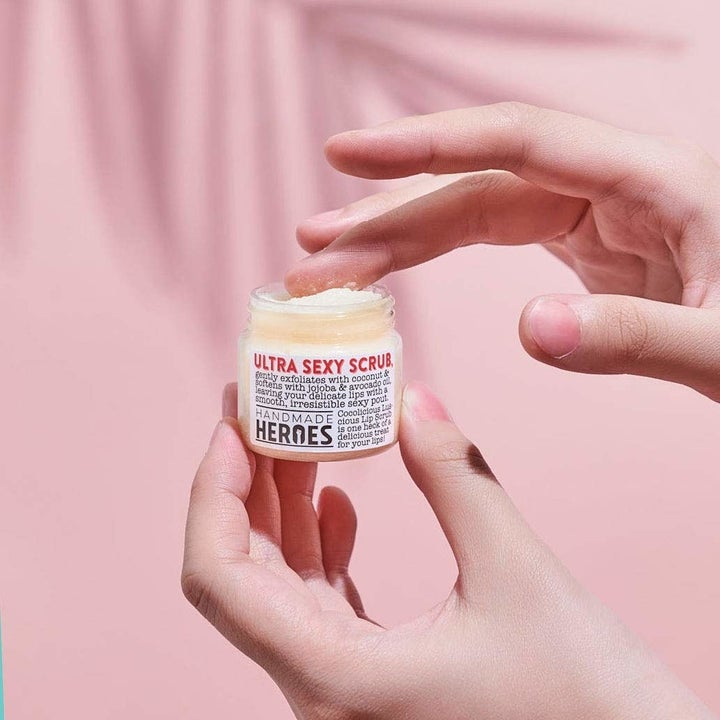 Finger scoops up cream-colored lip scrub from a tiny clear jar