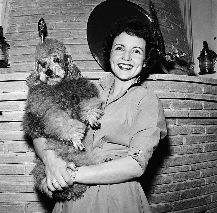 A young Betty White smiling while wearing a wide-brimmed hat, a shirt dress, and holding a dog