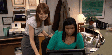 Two coworkers looking at a computer; one is slightly dancing happily while the other looks devoid of emotion