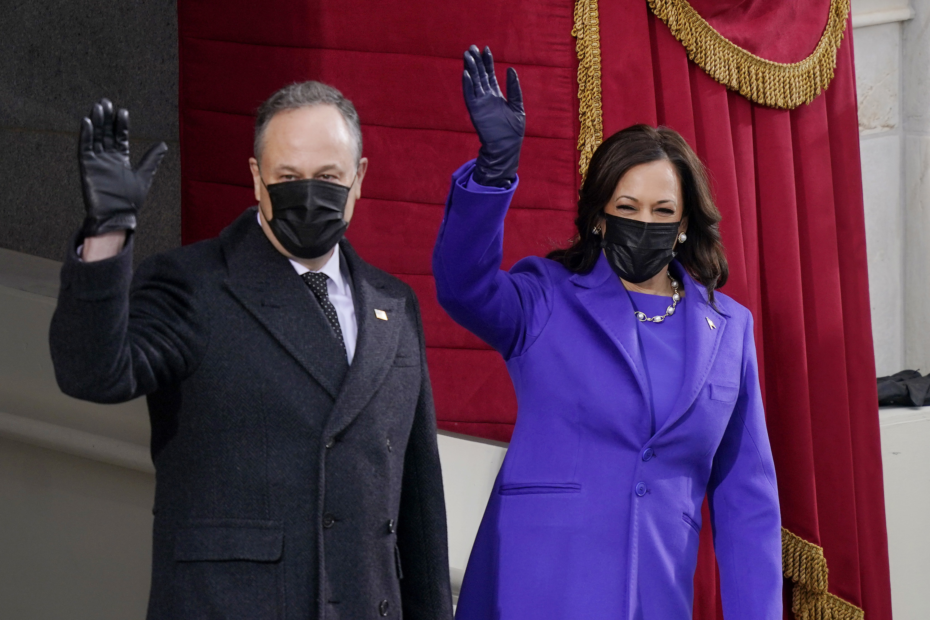 Vice President Kamala Harris in a purple suit and black face mask and her husband Doug Emhoff in a black face mask, standing and waving at the inauguration. 