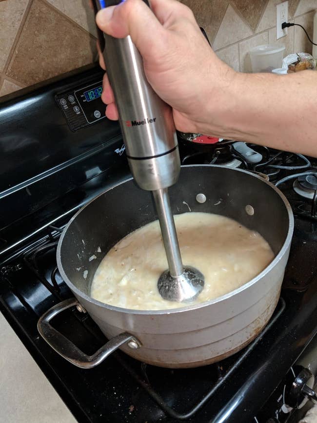 A reviewer photo of a hand using the immersion blender to blend a thick, white liquid in a pot