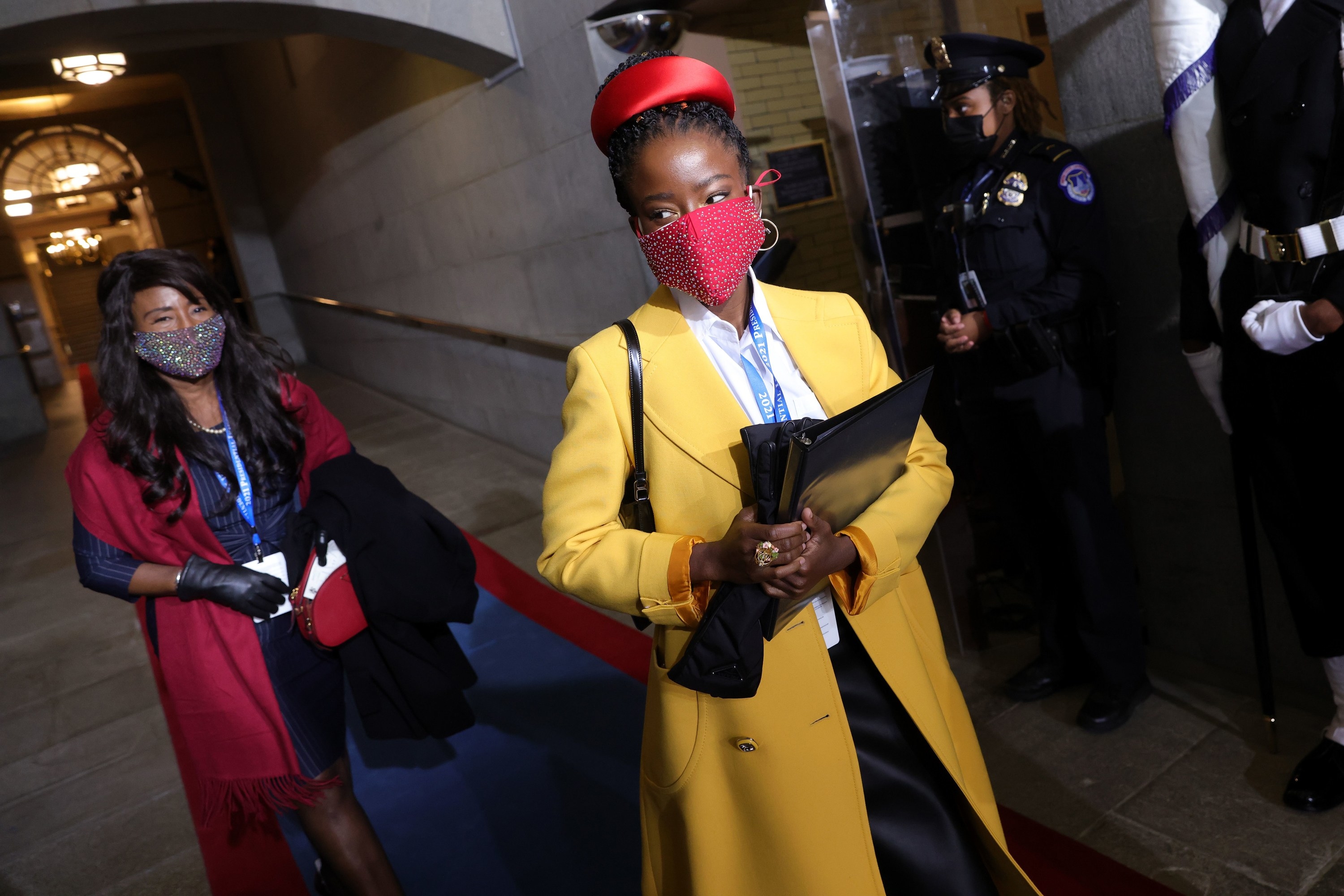 National youth poet laureate Amanda Gorman in a yellow coat and red studded face mask arriving at the inauguration, with another woman behind her in a red coat and floral face mask. 