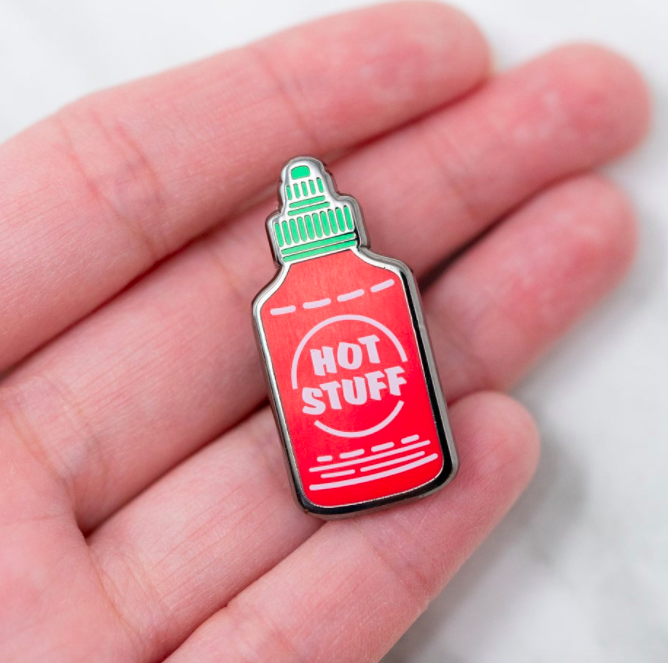 A person holding an hot sauce shaped enamel pin that says hot stuff