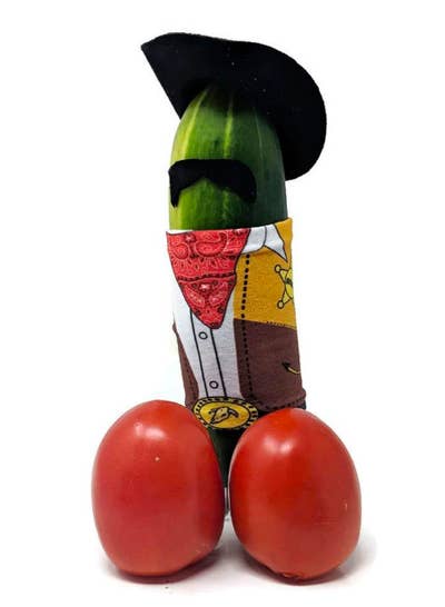 Cucumber with two cherry tomatoes in front of it. The cucumber is wearing a hat, mustache, and cowboy outfit. 
