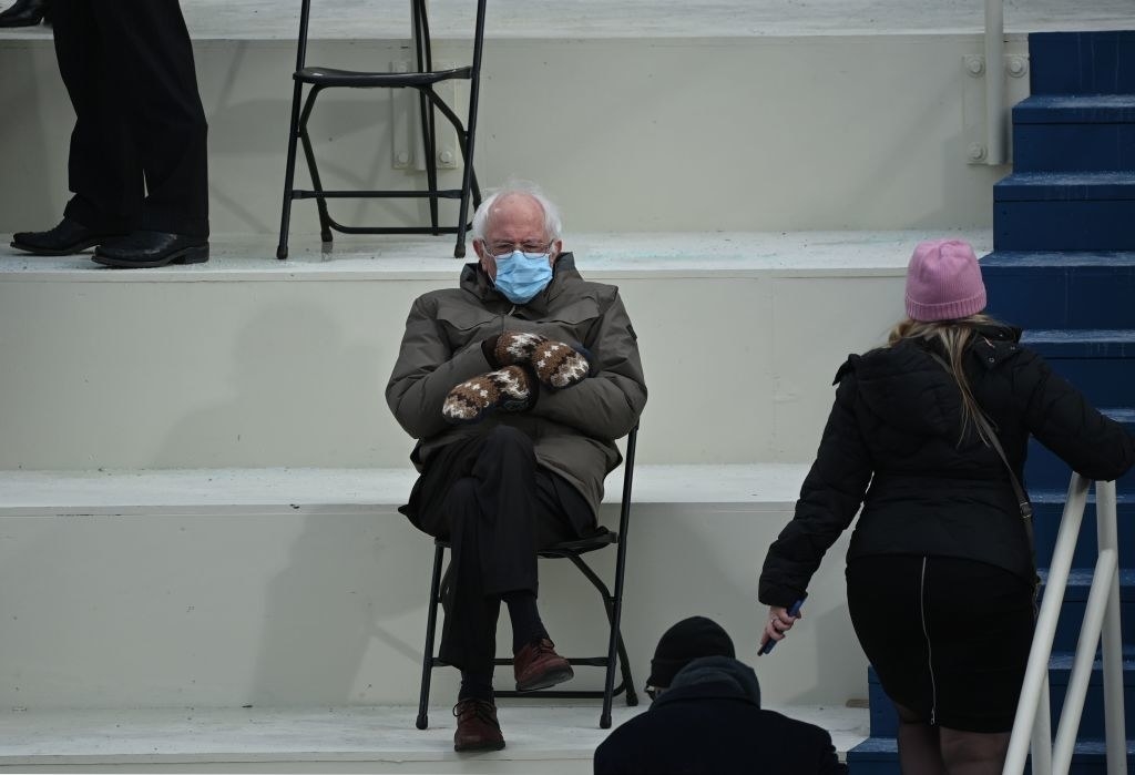 Bernie Sanders sitting on a folding chair wearing a coat, wool mittens, and a mask with his arms crossed