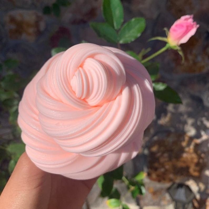 a hand holding a fluffy light pink slime next to a rose bush
