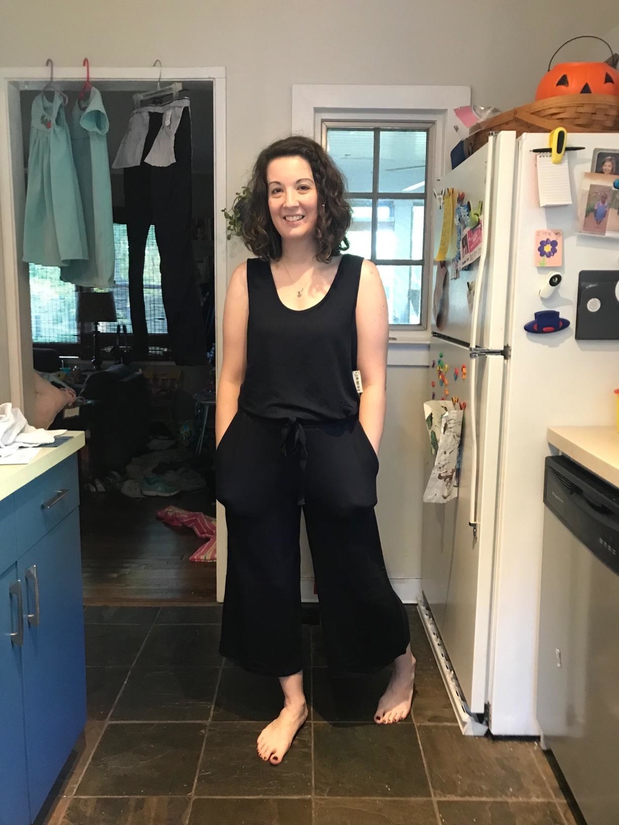 Reviewer wears black sleeveless terry-cloth jumpsuit while walking in their kitchen