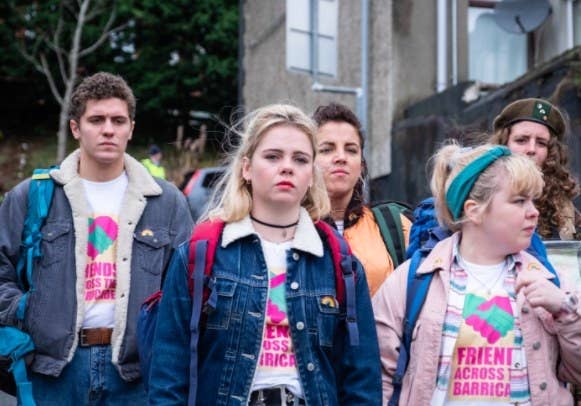 The cast of Derry Girls walk down the street, wearing &quot;friends across barricades&quot; tshirts and jackets 