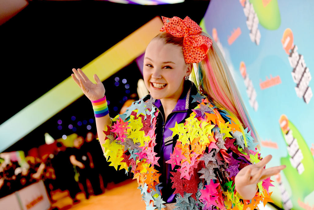 JoJo posing on a red carpet wearing a jacket decorated with cutout stars and a matching hair bow securing her signature side ponytail