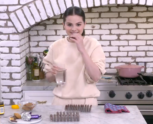 Selena in her kitchen, smiling at the camera; she is holding a Tim Tam and a glass of water