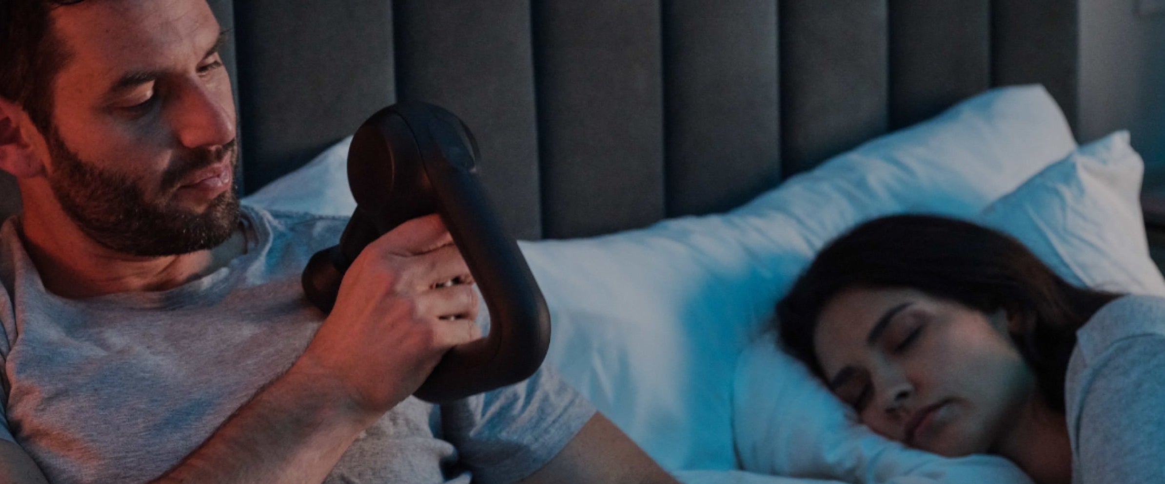 model holding the Theragun machine with a handle and ball at the end on their shoulder with another model asleep in bed next to them