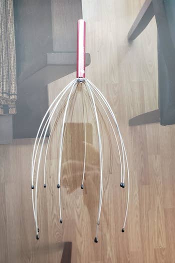 A reviewer photo of the scalp massage with a metallic pink handle and multiple prongs with rubber balls on the ends sitting on a table 