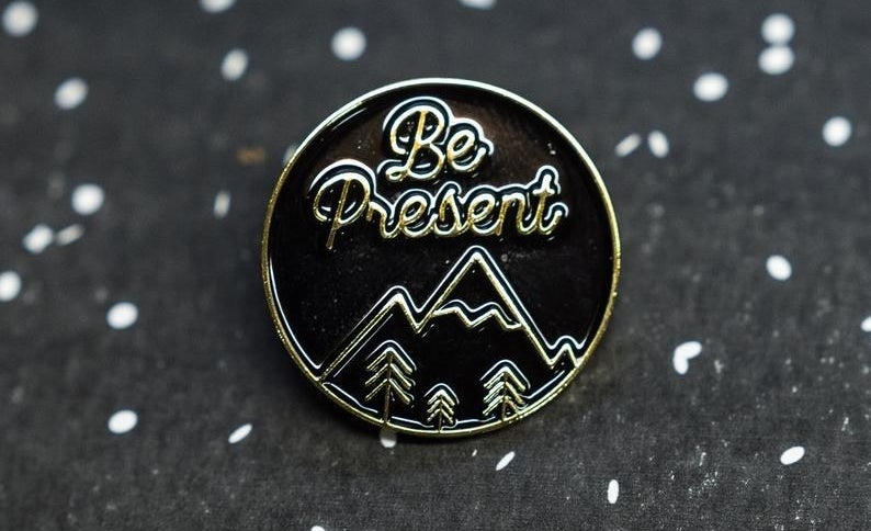 A round enamel pin with a black background and metallic mountainscape with text that reads &quot;Be Present&quot;