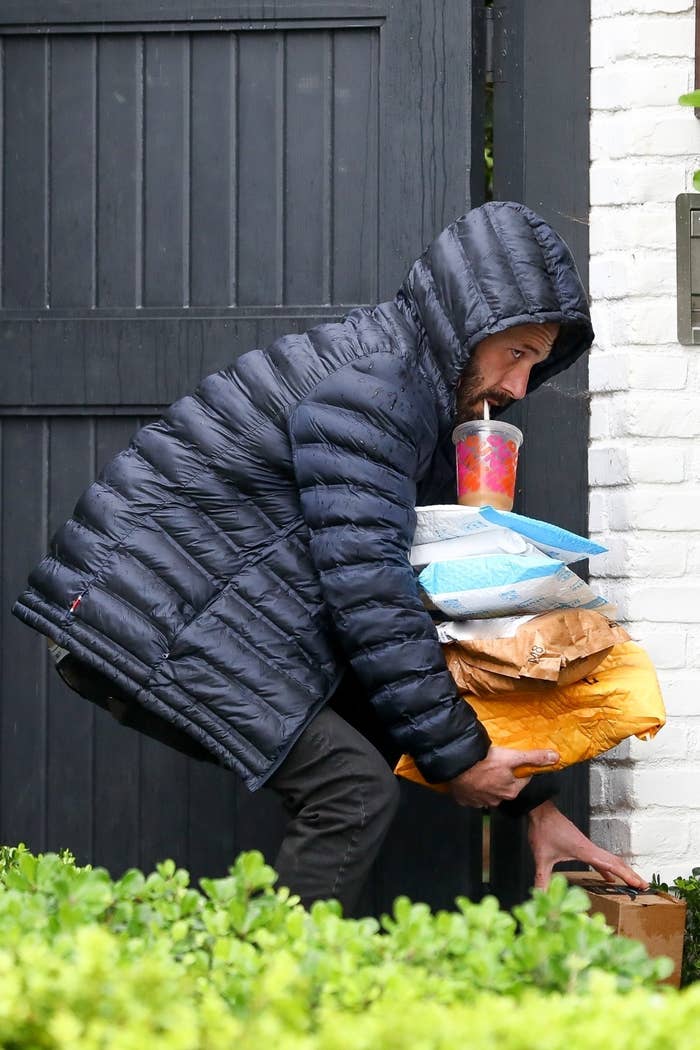 Ben Affleck, dressed in a puffy jacket with the hood up, hunches over to pick up a package with one hand as he balances several other packages with a large iced coffee sitting on top with his other arm