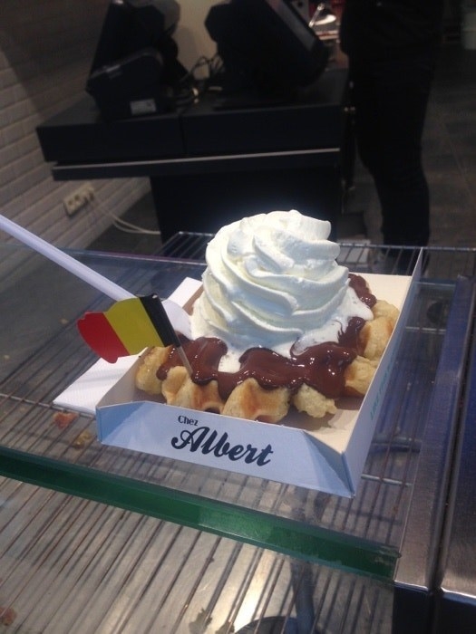 A small box holds a Belgian-style waffle topped with chocolate sauce, a pile of whipped cream, and a small Belgium flag on a toothpick