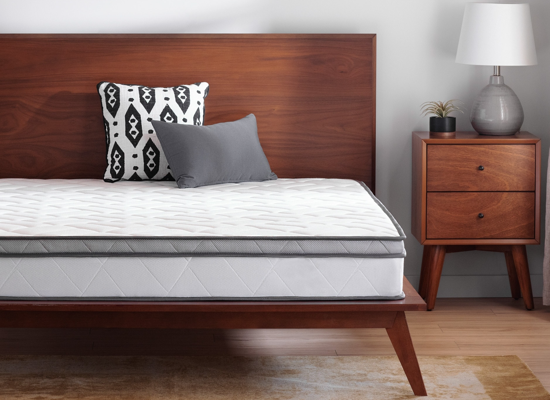 A mattress sitting on a bed frame. There is a nightstand by its side. There is a small plant and table lamp on the nightstand. 