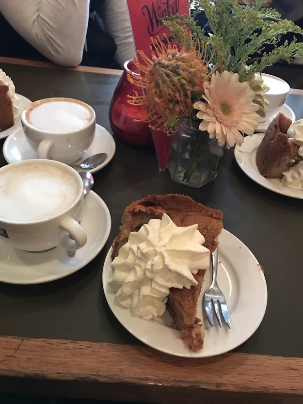 A plate with a large slice of pie topped with a ton of whipped cream sits on a table with coffee drinks and a vase of flowers