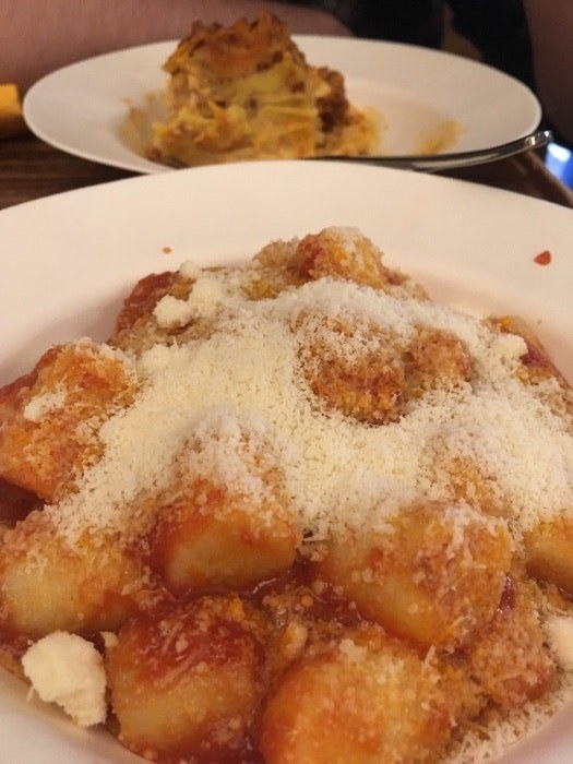 A plate of gnocchi in red sauce and covered in grated cheese in the foreground, with another dish in the blurred background