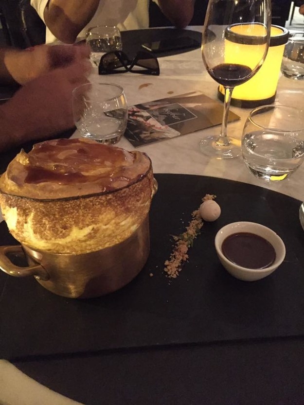 A soufflé expands out of a small metal pot sitting on a table next to a small bowl of sauce