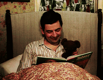 Mr Bean happily enjoying a book in bed with his stuffed animal