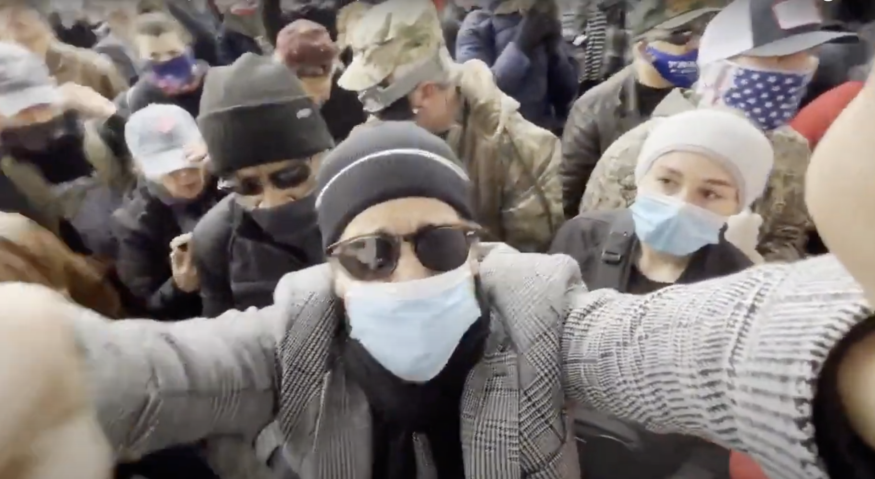 In a video screenshot, Brandon Straka, wearing a beanie, sunglasses, face mask, and jacket, looks into the camera amid a crowd of people