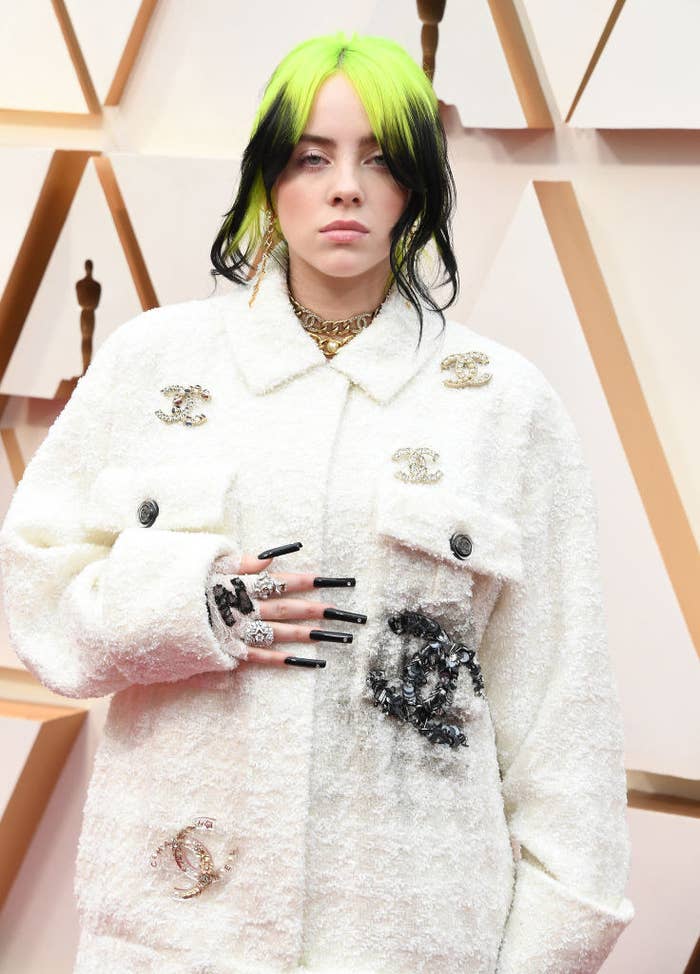 Billie Eilish posing on a red carpet wearing matching Chanel jacket and pants featuring several brooches shaped into the iconic interlocking double C