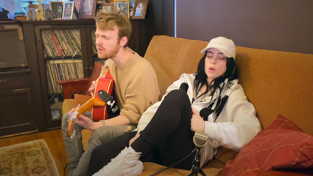 Billie and Finneas, who&#x27;s playing a guitar, sitting on a couch recording a song