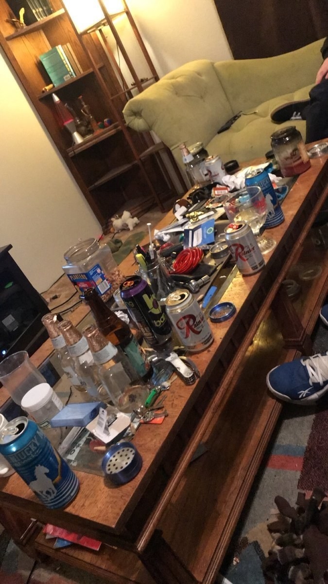 The coffee table has old beer bottles, beer cans, glasses, half-empty jars of snacks and general dirt all over it
