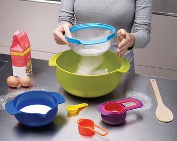 A model using a strainer over a large green bowl surrounded by a blue bowl and several different-colored measuring cups 