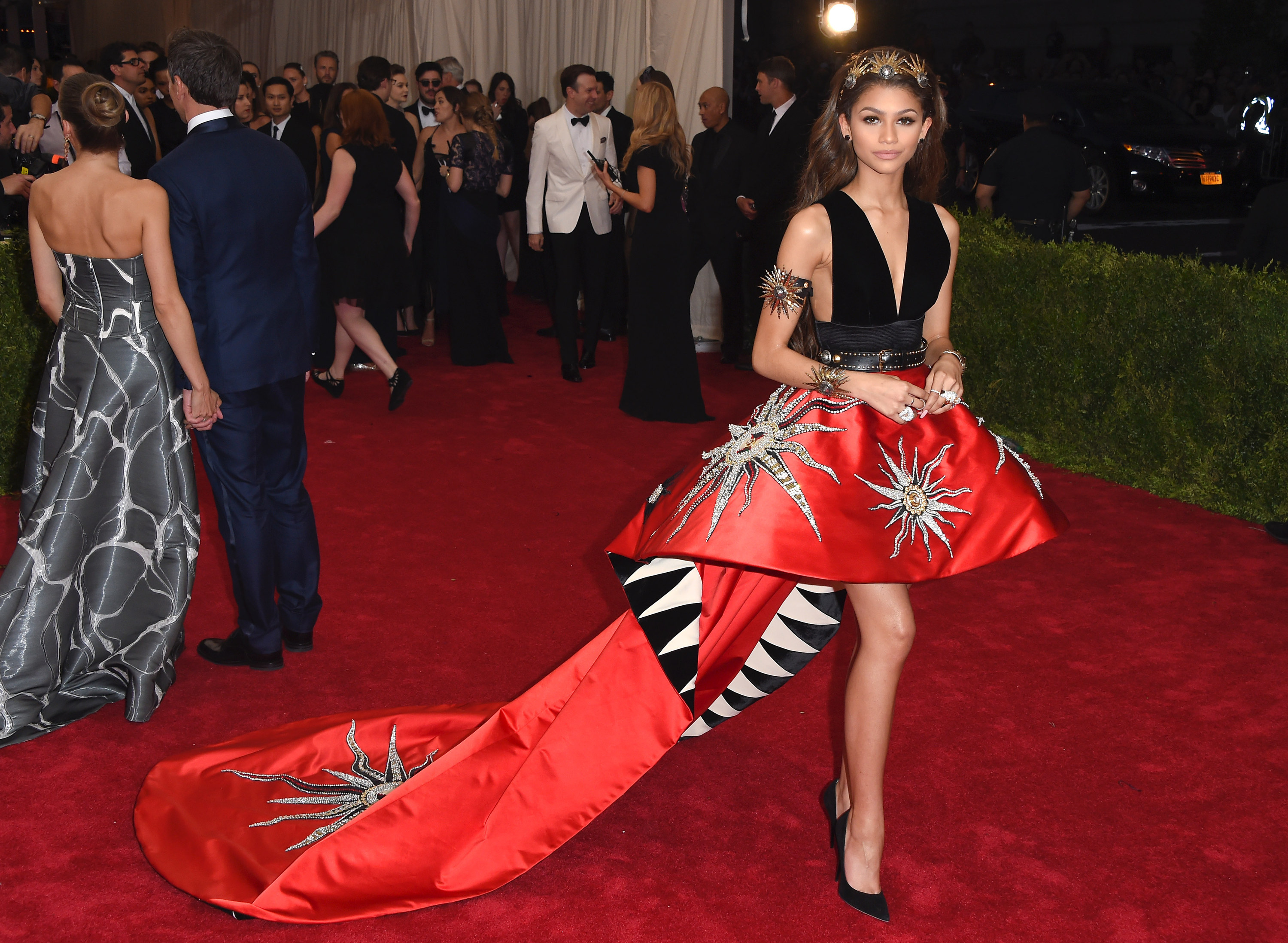 Zendaya wearing a dress with a long train covered in artistically drawn suns, she&#x27;s also wearing a crown on her head. 