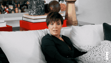 Kris Jenner sitting on a couch while wearing very long earrings seemingly staring into space