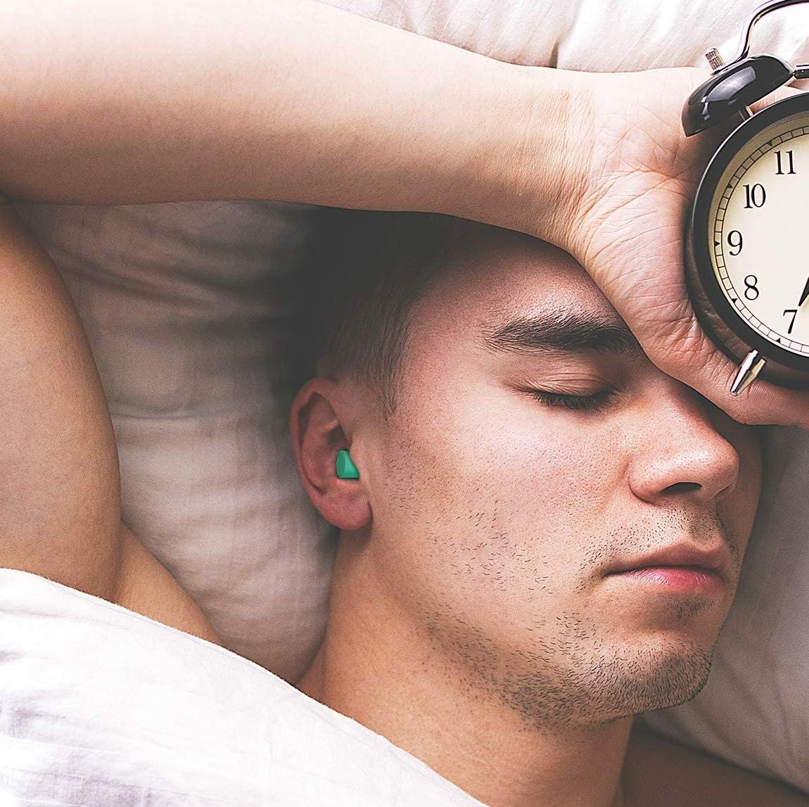 person lying in bed with earplugs in holding an alarm clock