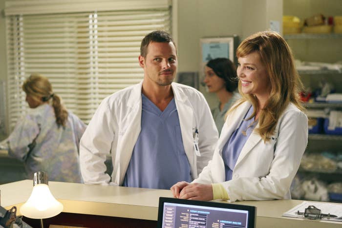 Justin Chambers, wearing a white lab coat and a blue shirt, stands with his hands on his shoulders next to Katherine Heigl, leaning on a counter while wearing the same, in Grey&#x27;s Anatomy