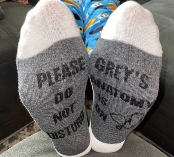 reviewer wearing the socks in gray and white — one says &quot;please do not disturb&quot; in black text and the other says &quot;grey&#x27;s anatomy is on&quot; in black text with an illustration of a stethoscope