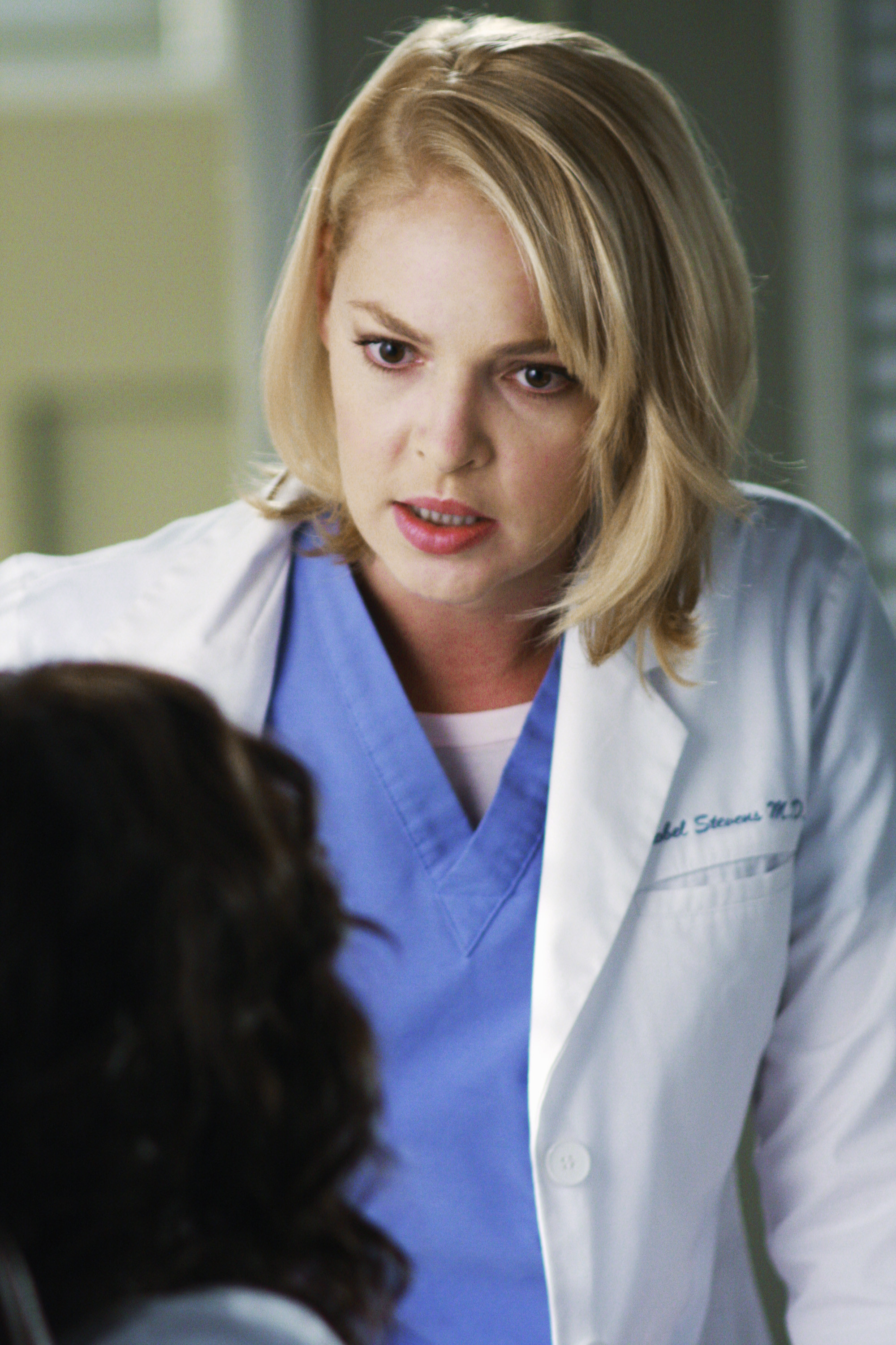 Katherine Heigl, as Izzie Stevens, wears a white lab coat and blue scrubs while talking to a patient in Grey&#x27;s Anatomy