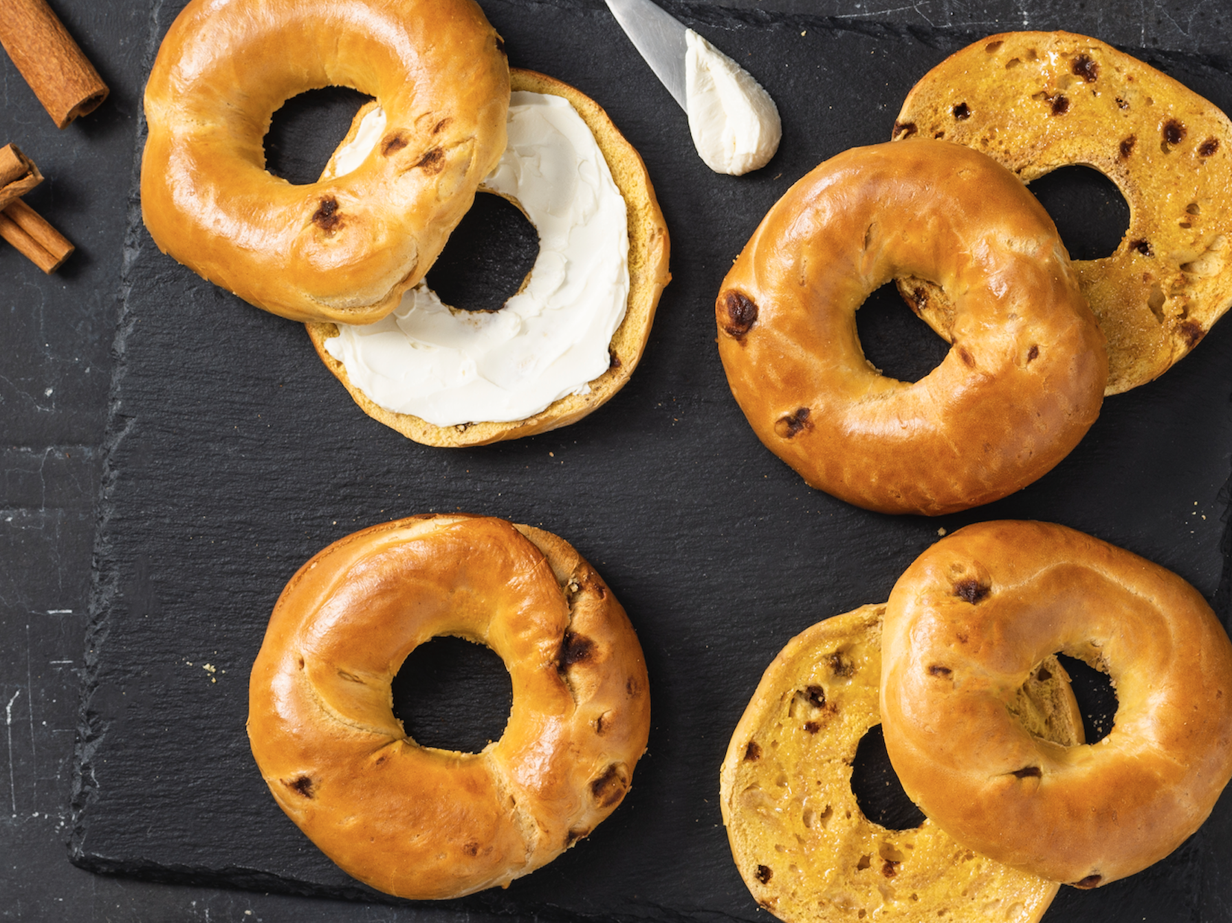 An overhead photo of Maple French Toast bagels on black slate. Some are cut open and some have cream cheese spread on top.