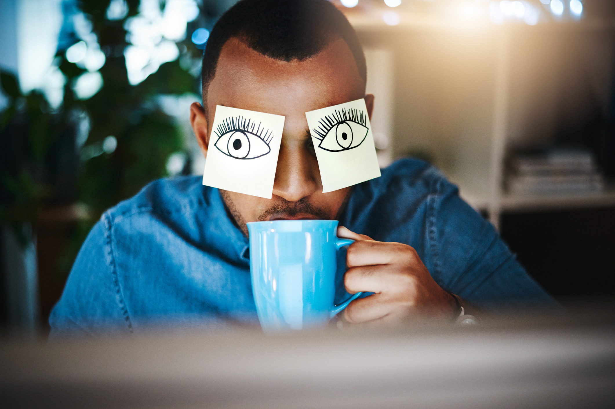 A man sitting in front of a computer drinking from a coffee mug. Two pieces of papers are over his eyes with drawn eyes on top.