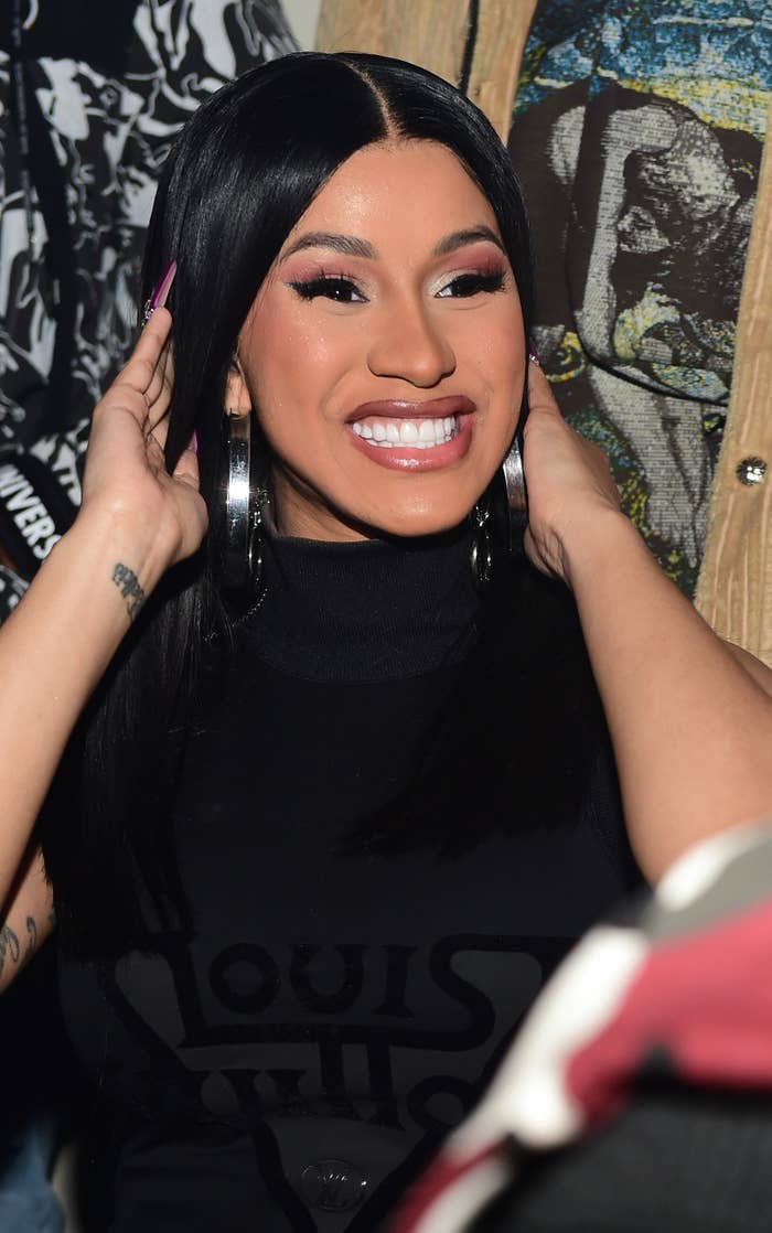 Cardi B, wearing a dress and sporting long straight hair and long nails, smiles at a party