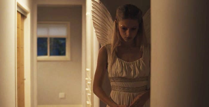 A blonde girl wearing angel wings stands in a hallway looking down