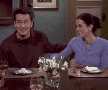 Chandler and Monica fake laughing at a table pretending to be enthused but ultimately having each other&#x27;s backs in this particular awkward moment at Chandler&#x27;s boss&#x27;s house