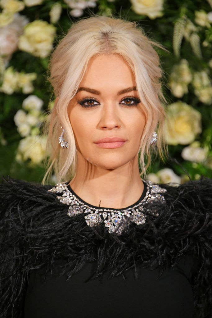Rita Ora posing at an event wearing a dress with a feathered and bejeweled collar, drop earrings, and her hair in an updo