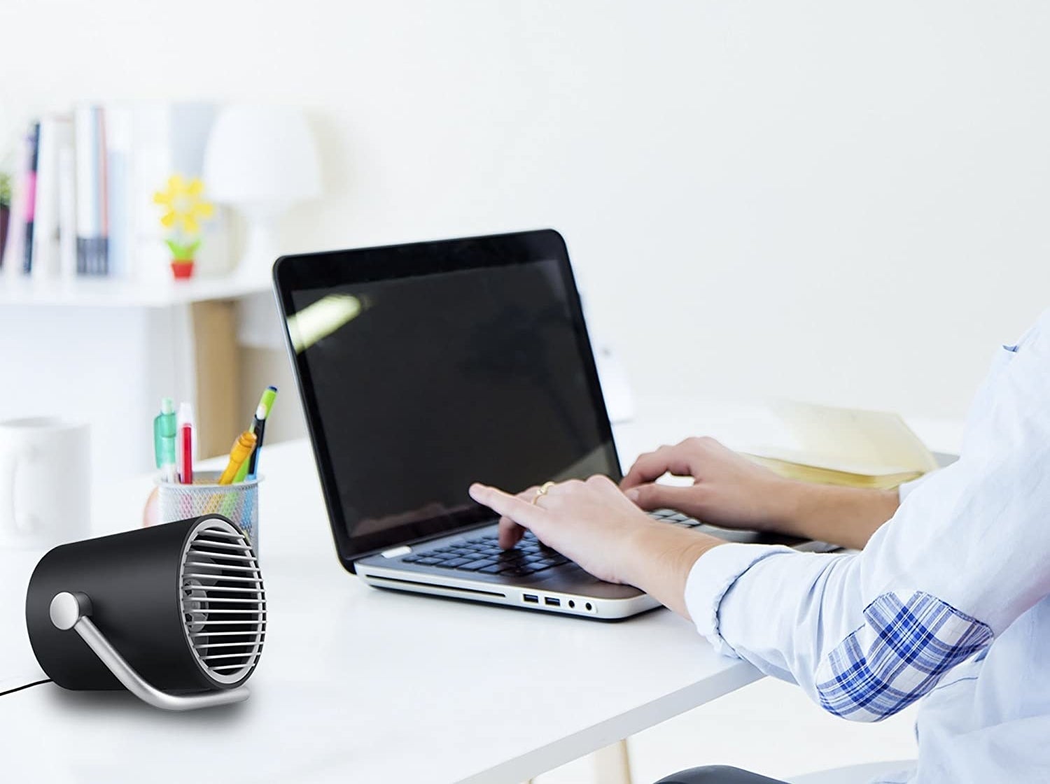 A person sitting at a desk working on a laptop beside the small fan